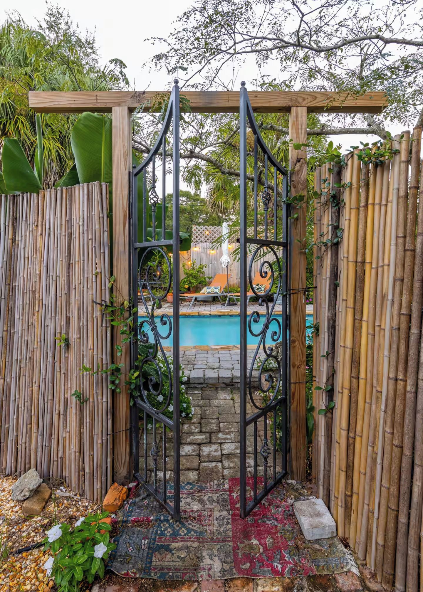 A gated entrance to a pool area with a bamboo fence, a metal gate, and a patterned rug on the ground.