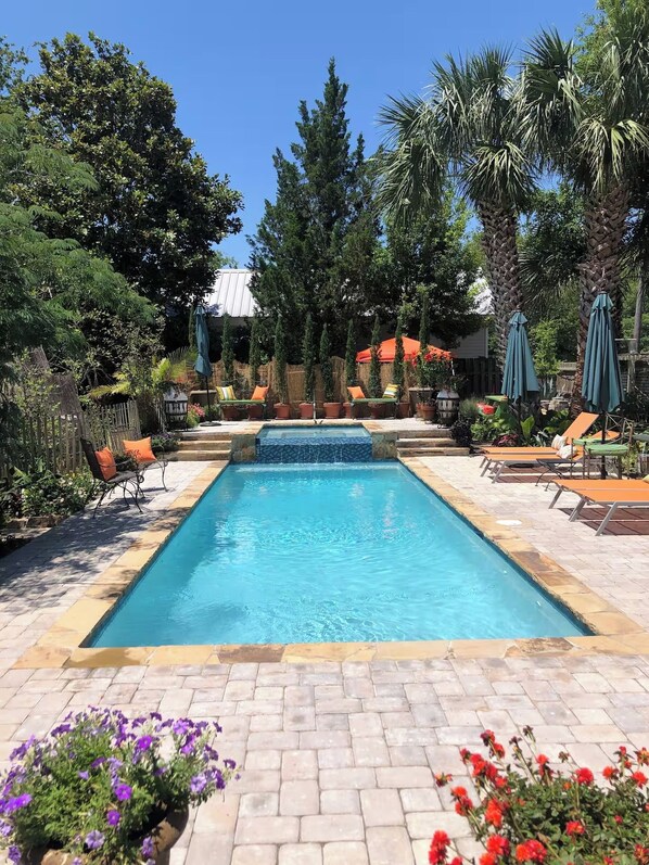 Una piscina rectangular con borde de azulejos, rodeada de sillas de descanso y sombrillas, con árboles y una valla al fondo.