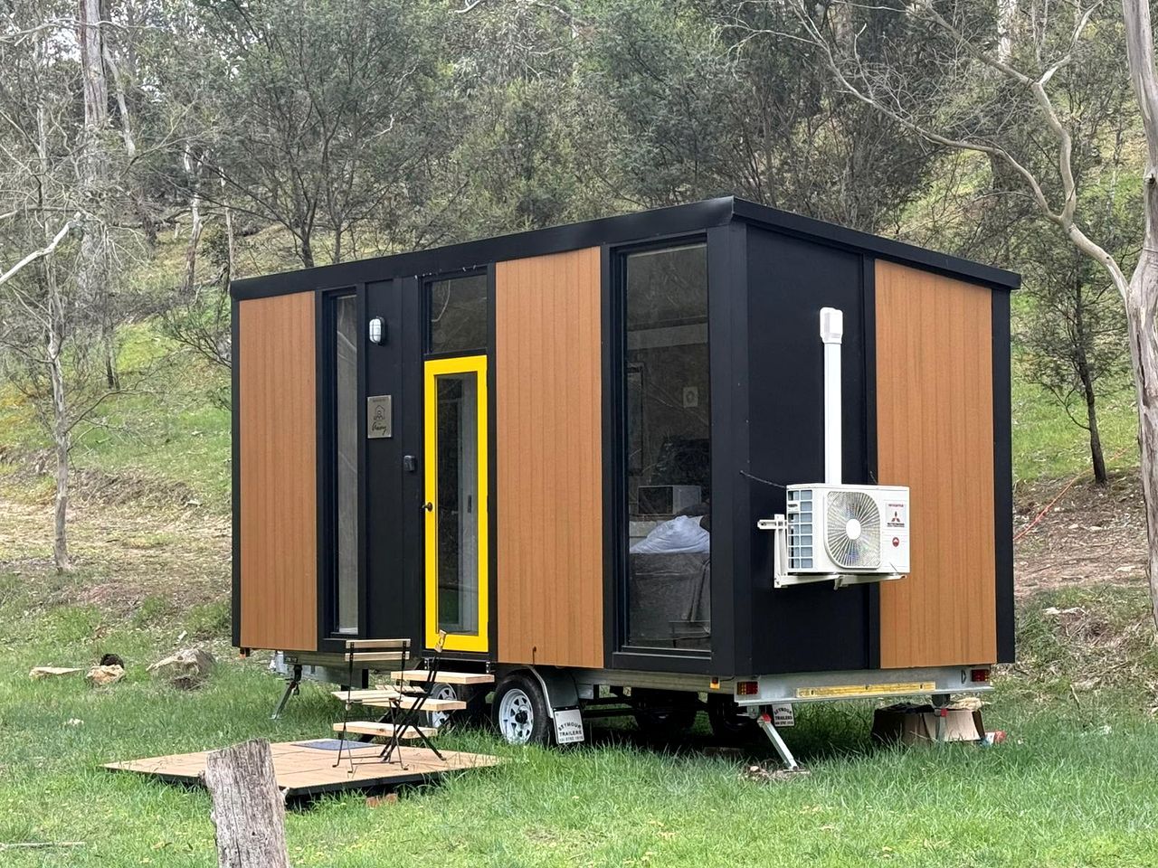 A small, modern mobile home with a sliding door, an air conditioning unit, and a portable table.