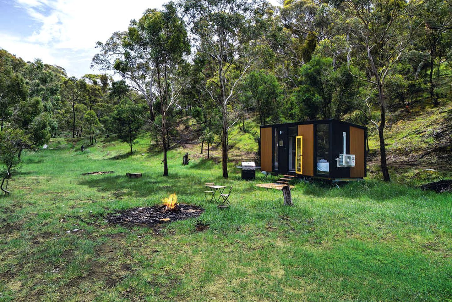 Eine kleine Holzhütte mit Feuerstelle in einer grünen, von Bäumen umgebenen Wiese.