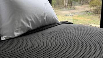 Un lit avec un oreiller blanc et une couverture grise à texture, une fenêtre donnant sur des arbres et une zone herbeuse.