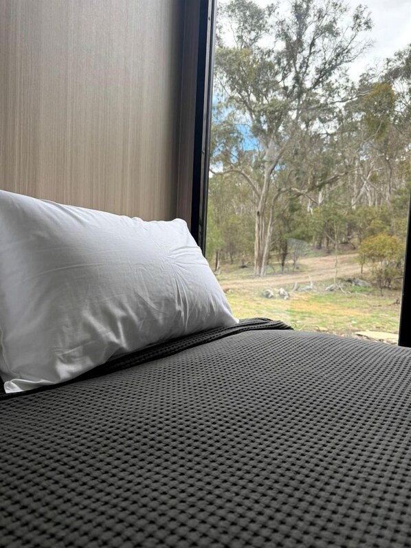 A bed with a white pillow and a grey textured bedspread, a window with a view of trees and a grassy area.