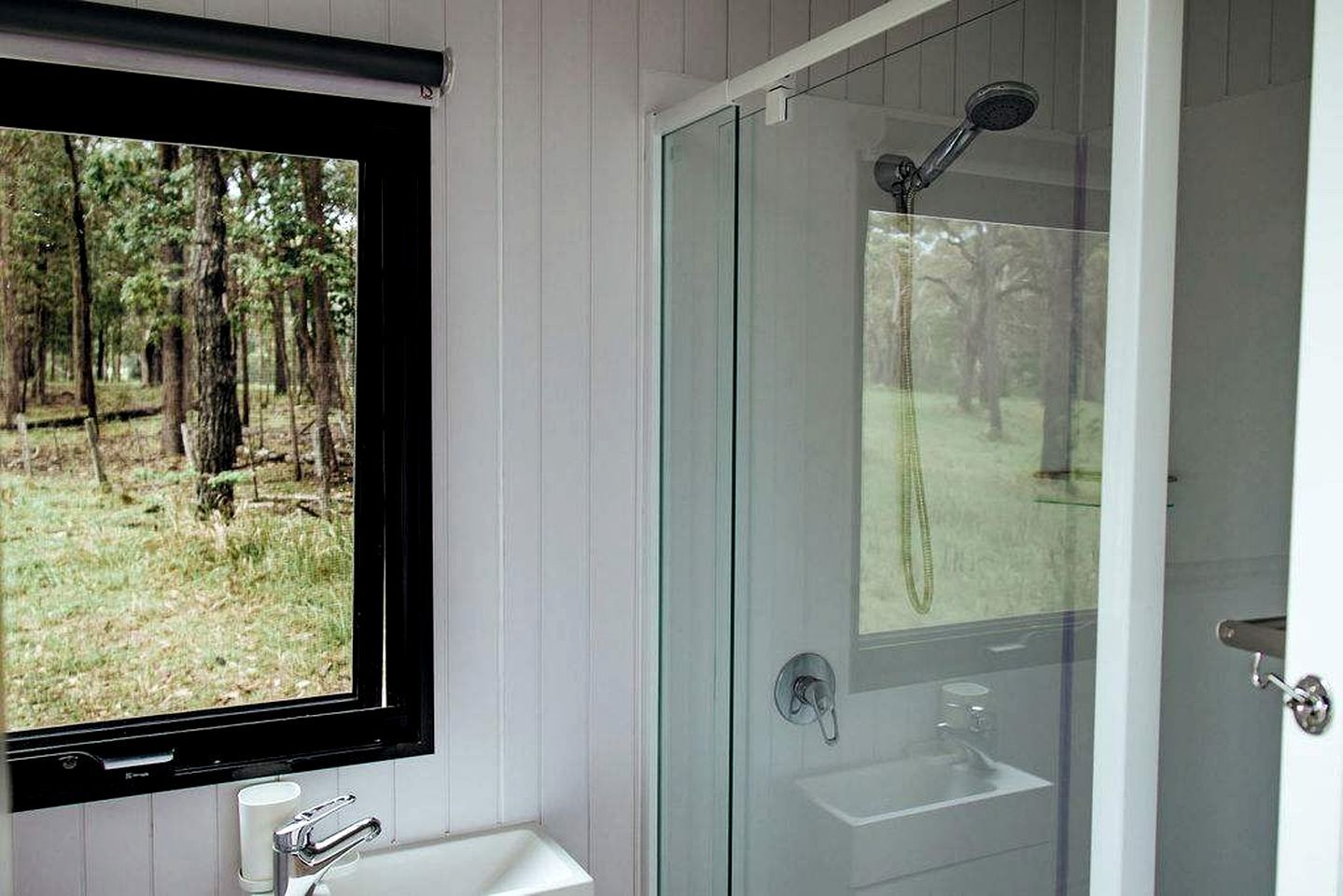 A bathroom with a glass shower enclosure, a sink, and a window overlooking a forest.