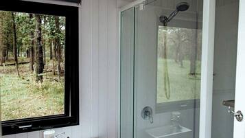 A bathroom with a glass shower enclosure, a sink, and a window overlooking a forest.
