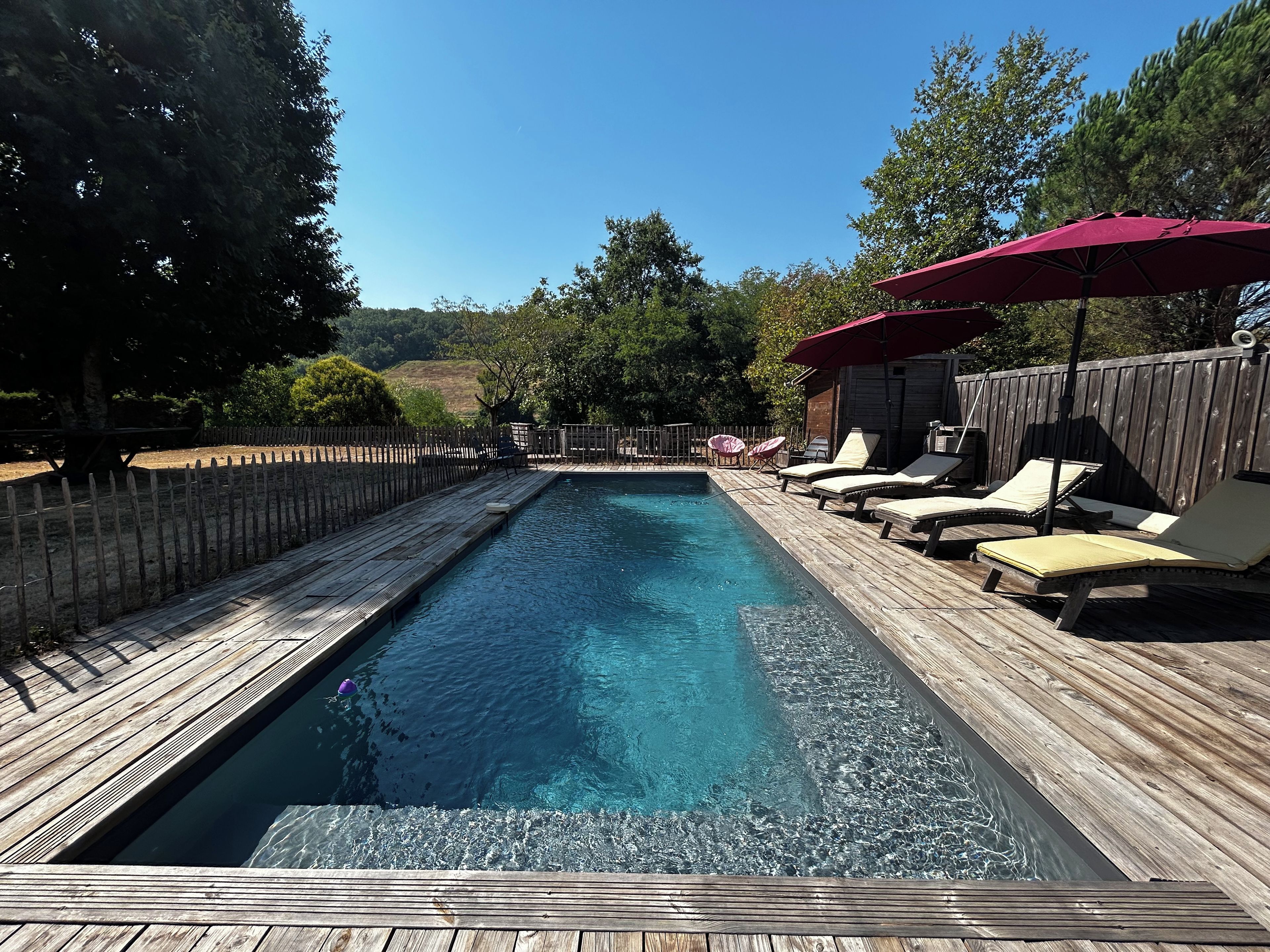 A rectangular swimming pool with a wooden deck, surrounded by lounge chairs and a red umbrella.