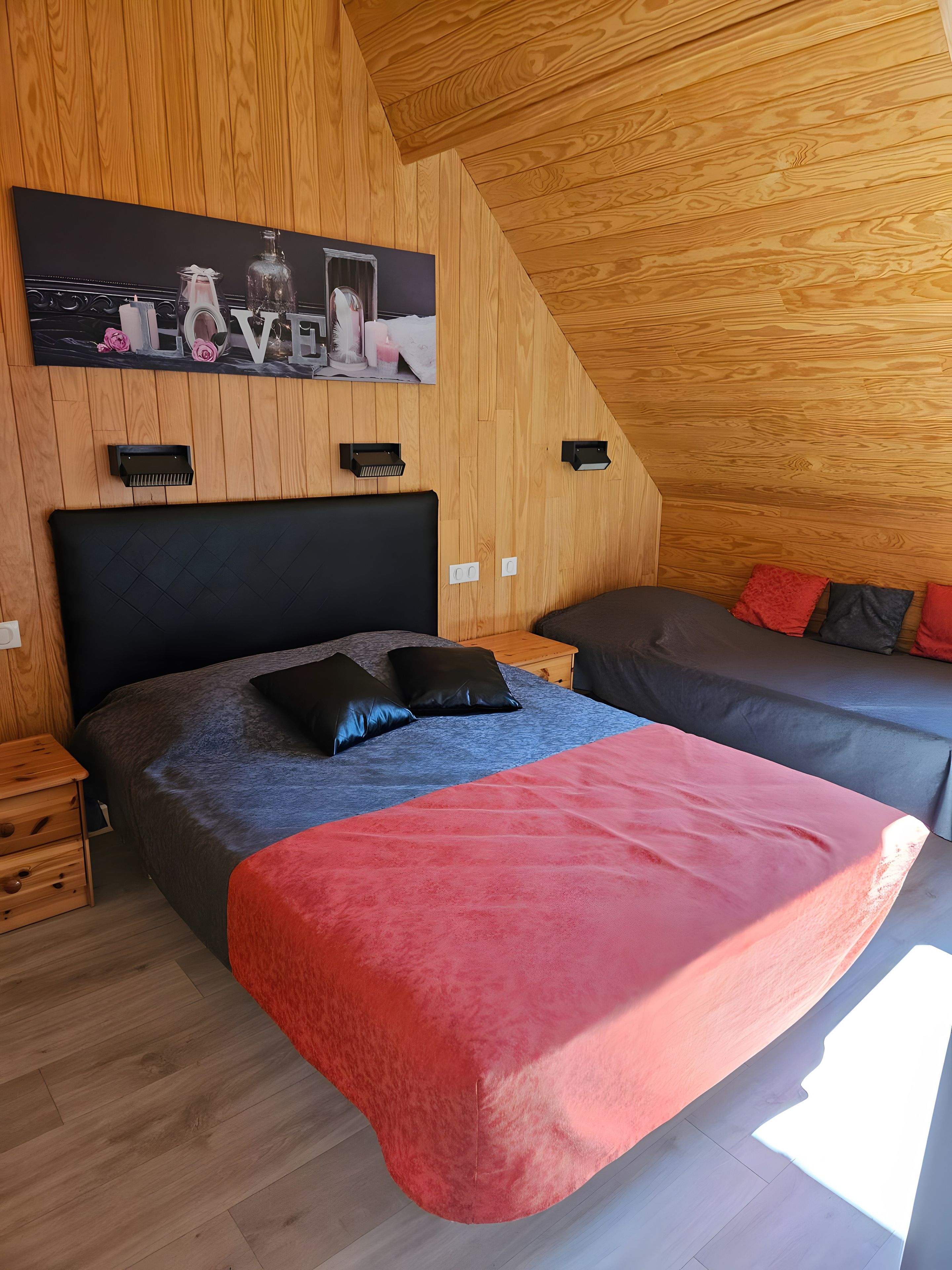 Ein Schlafzimmer mit schrägem Dach, einem Bett mit roter Decke, einem grauen Sofa und einem Gemälde an der Wand.