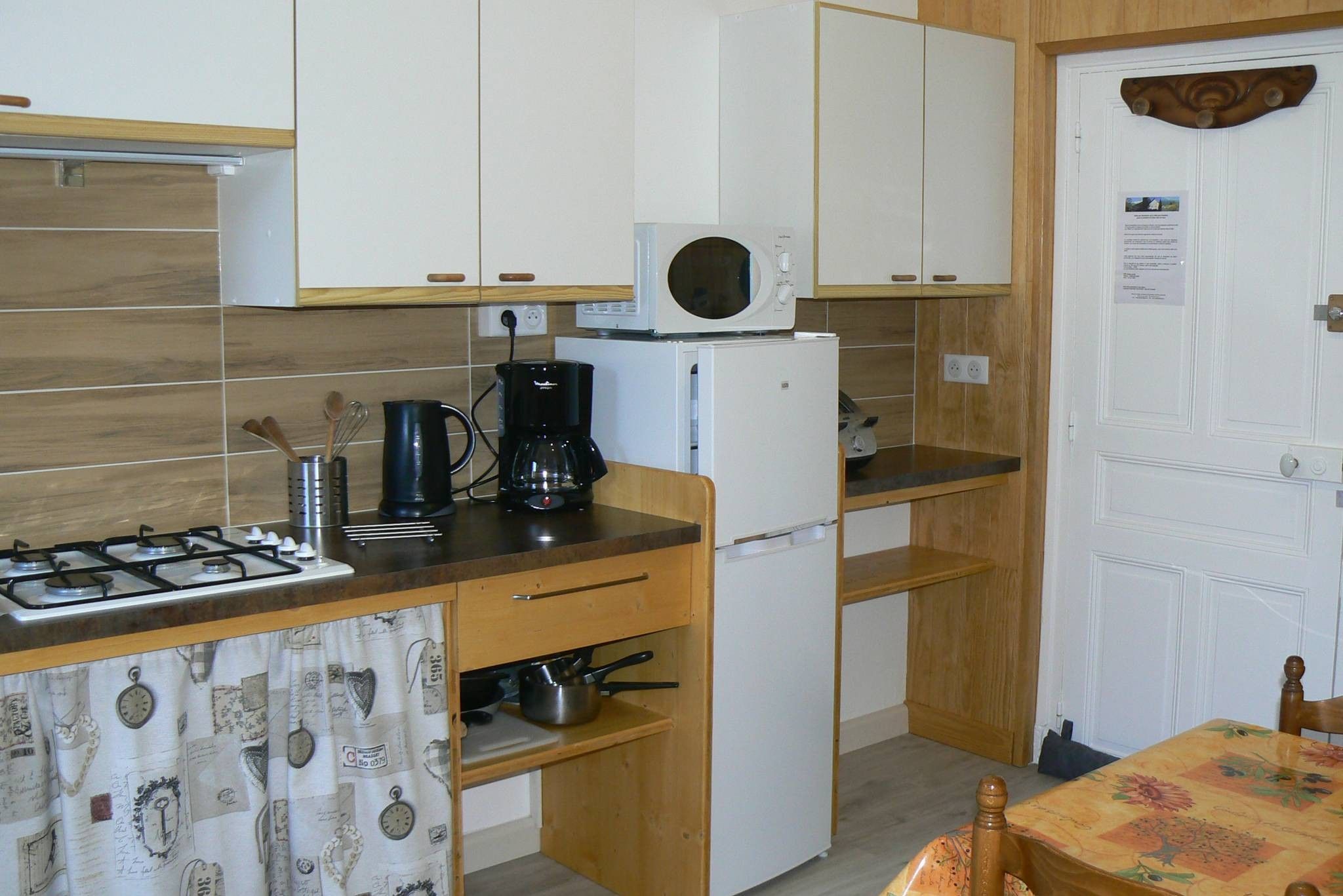 A compact kitchen with a gas stove, a microwave, a coffee maker, and a refrigerator. There is a wooden table with a floral tablecloth and a chair.
