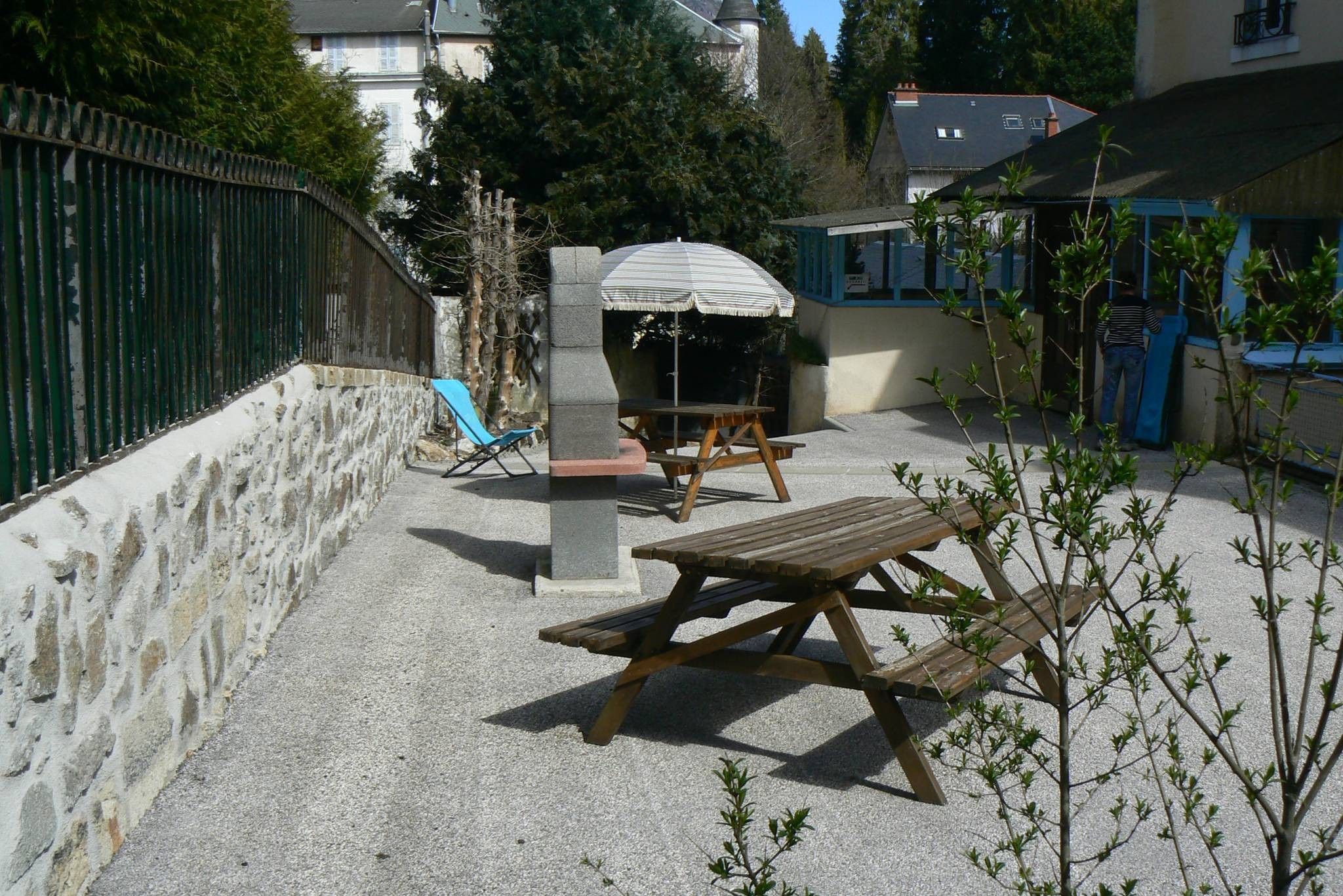 An outdoor patio area with a stone wall, a wooden table, a blue chair, and a picnic table with a striped umbrella.