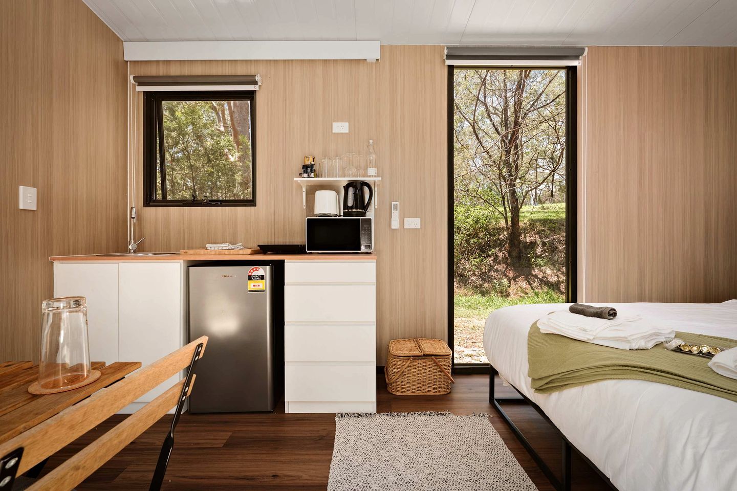 Habitació compacte amb un llit, una cuineta petita, un refrigerador i una finestra amb vista a arbres.