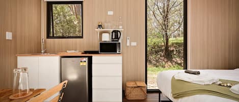 Habitació compacte amb un llit, una cuineta petita, un refrigerador i una finestra amb vista a arbres.