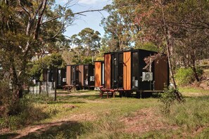 A row of modern, compact cabins with external air conditioning units, situated in a forested area with a dirt path leading to them. - Cozy Holiday Home with Eco Design and Firepit Escape in Maroota, Australia (Canoelands)