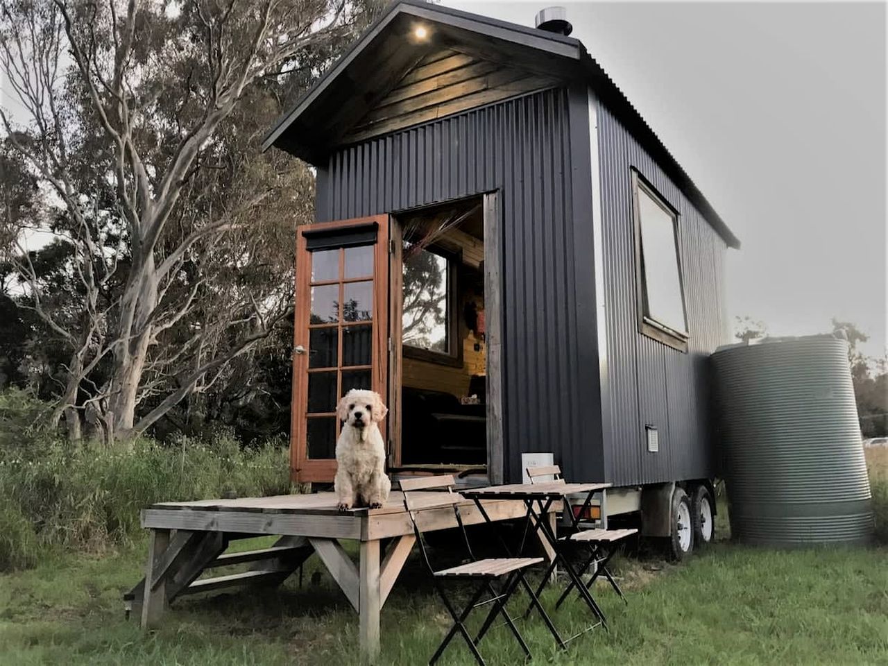 A small, dark-colored cabin with a wooden door and a dog sitting outside.