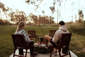 Two people seated at a round table with a fire pit in the center, outdoors with trees in the background.