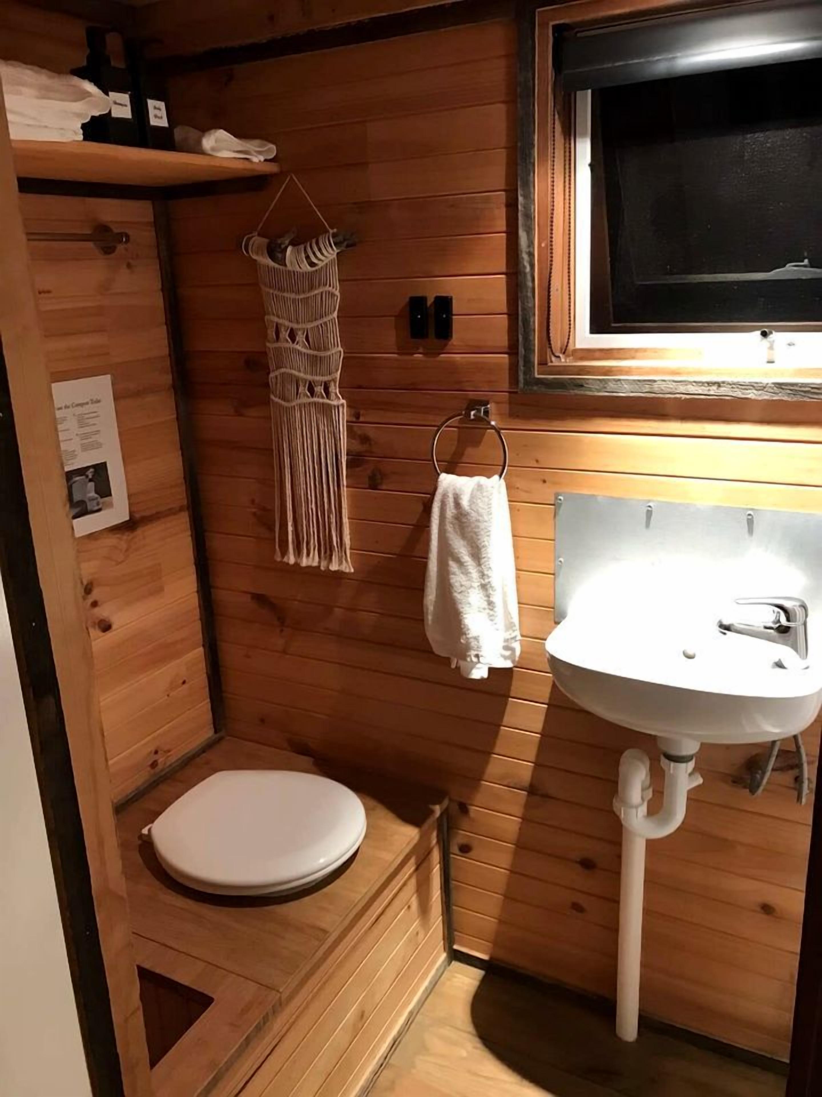 A compact wooden bathroom with a white toilet, a sink, and a macramé towel rack.