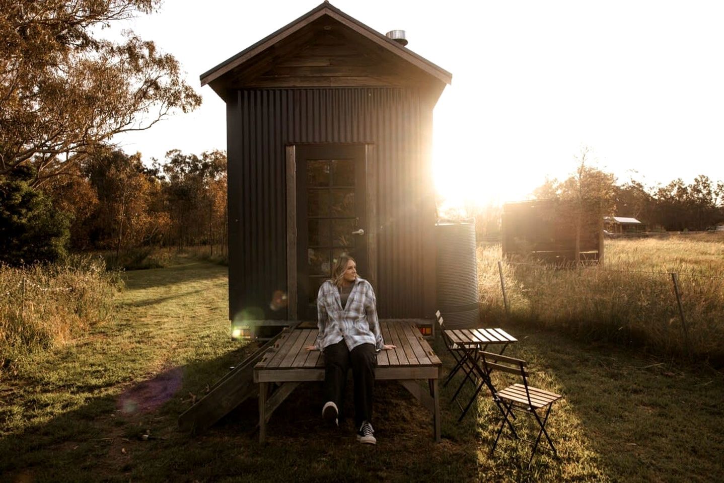Une personne est assise sur une terrasse en bois devant une petite construction en bois dotée d’un toit en pente, et l’ensemble est entouré d’herbe et d’arbres.