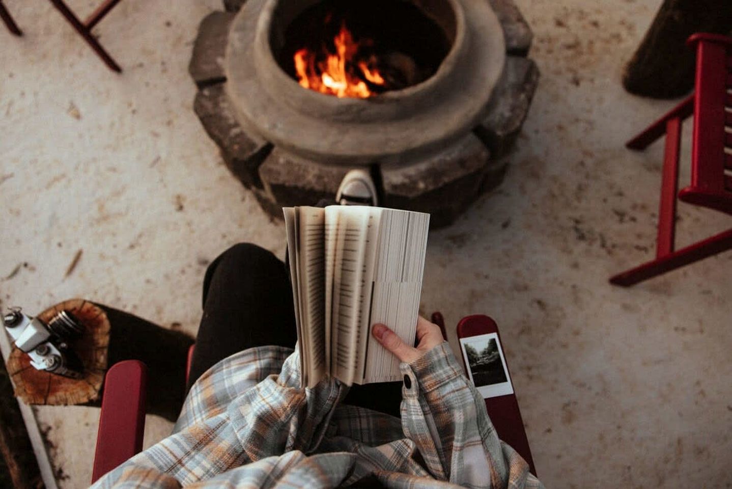 A person sitting in a chair with a book, a fire pit in the background, and a red chair to the side.