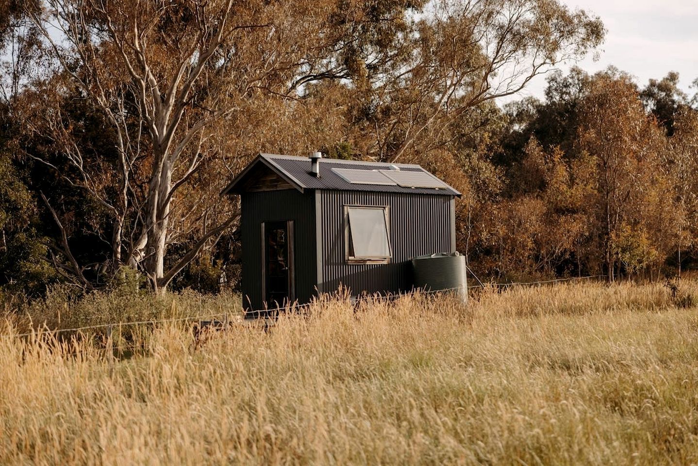 Een klein, donker houten huisje met een schuin dak, gelegen in een grasveld met bomen op de achtergrond.
