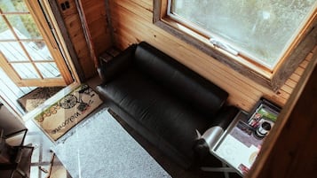 Una habitación pequeña con un sofá negro, una mesita y una ventana que da a un paisaje con árboles.