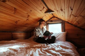 A person sitting on a bed in a wooden room with a sloped ceiling and a window. - Yarra Valley Tiny House for Eco-Friendly Glamping in Victoria (Yea)