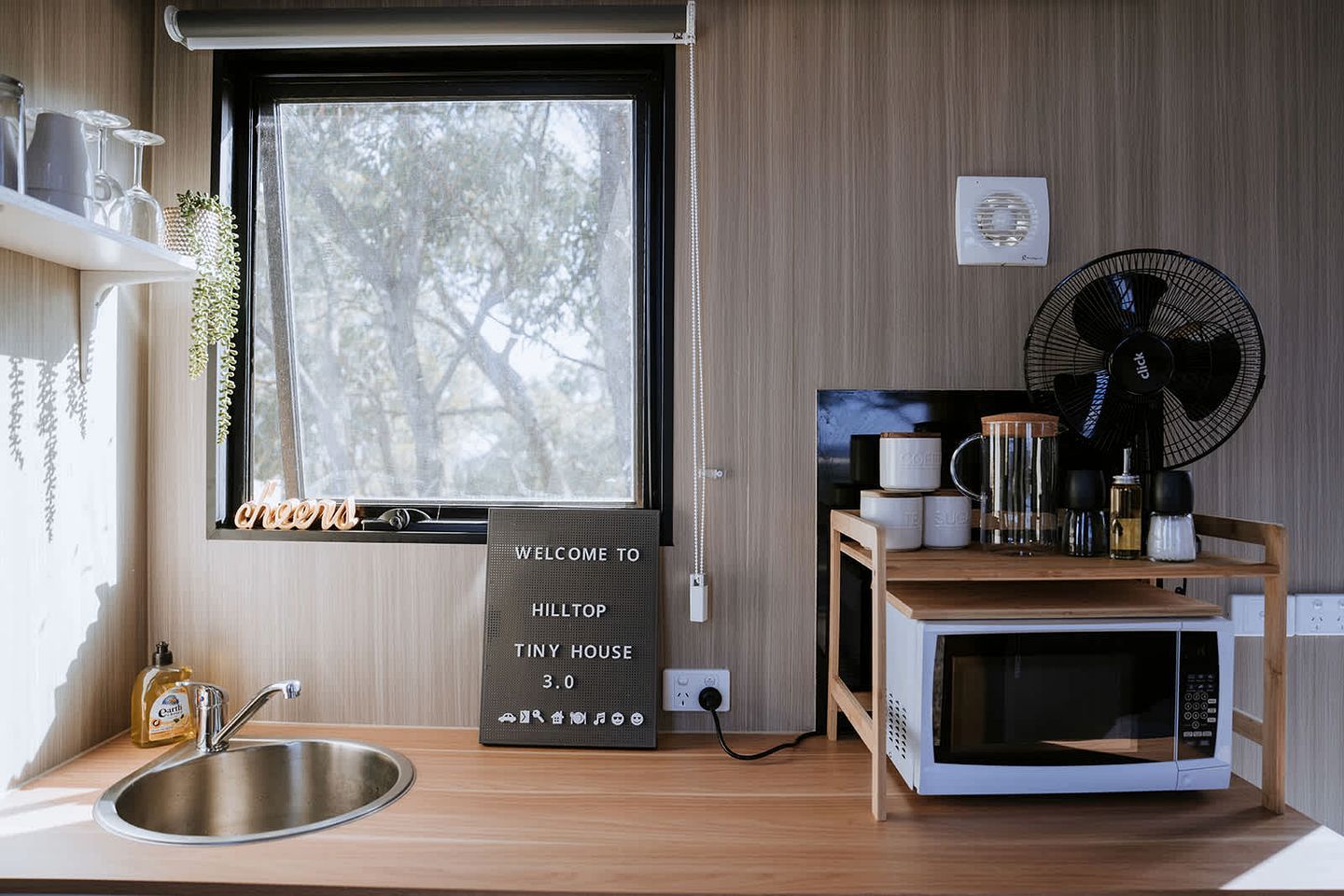Een kleine keuken met een spoelbak, een magnetron, een ventilator en een bord dat verblijf in Hilltop Tiny House welkom heet.