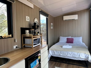 A hotel room with a bed, a small kitchenette, a microwave, a fan, and an air conditioning unit.