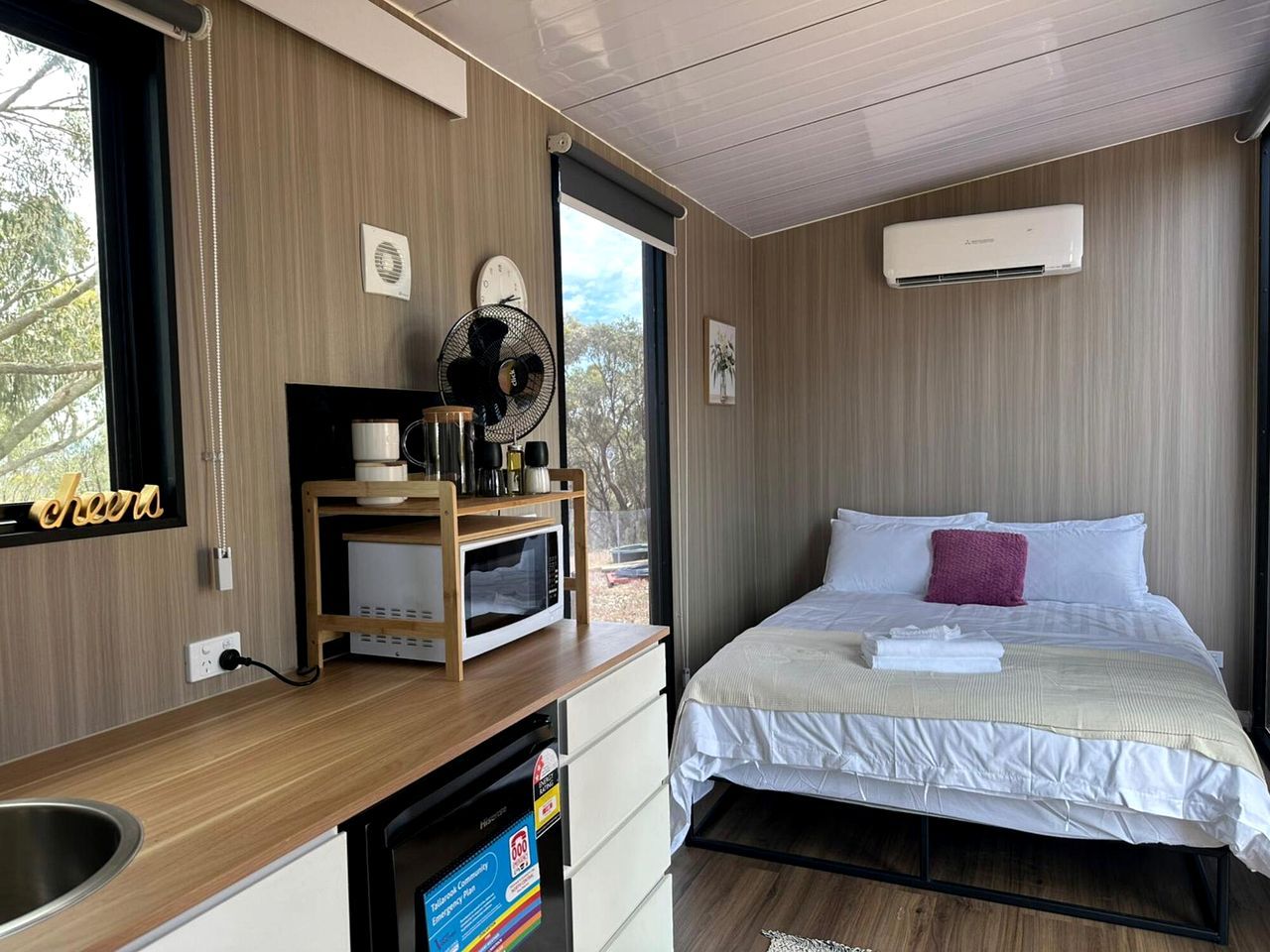 A hotel room with a bed, a small kitchenette, a microwave, a fan, and an air conditioning unit.