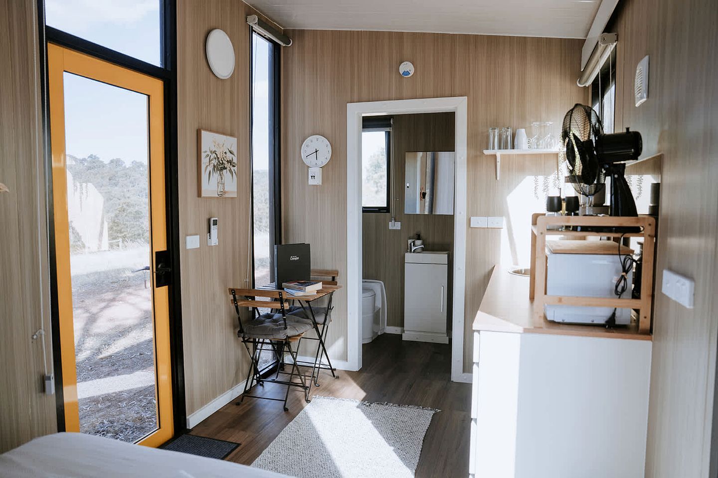 Et kompakt, moderne køkken med træskabe, et spiseområde med bord og stole, samt et synligt ur på væggen.