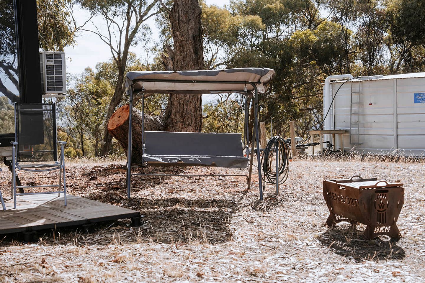 A rustic outdoor seating area with a metal bench and a covered table, surrounded by trees and dry grass.