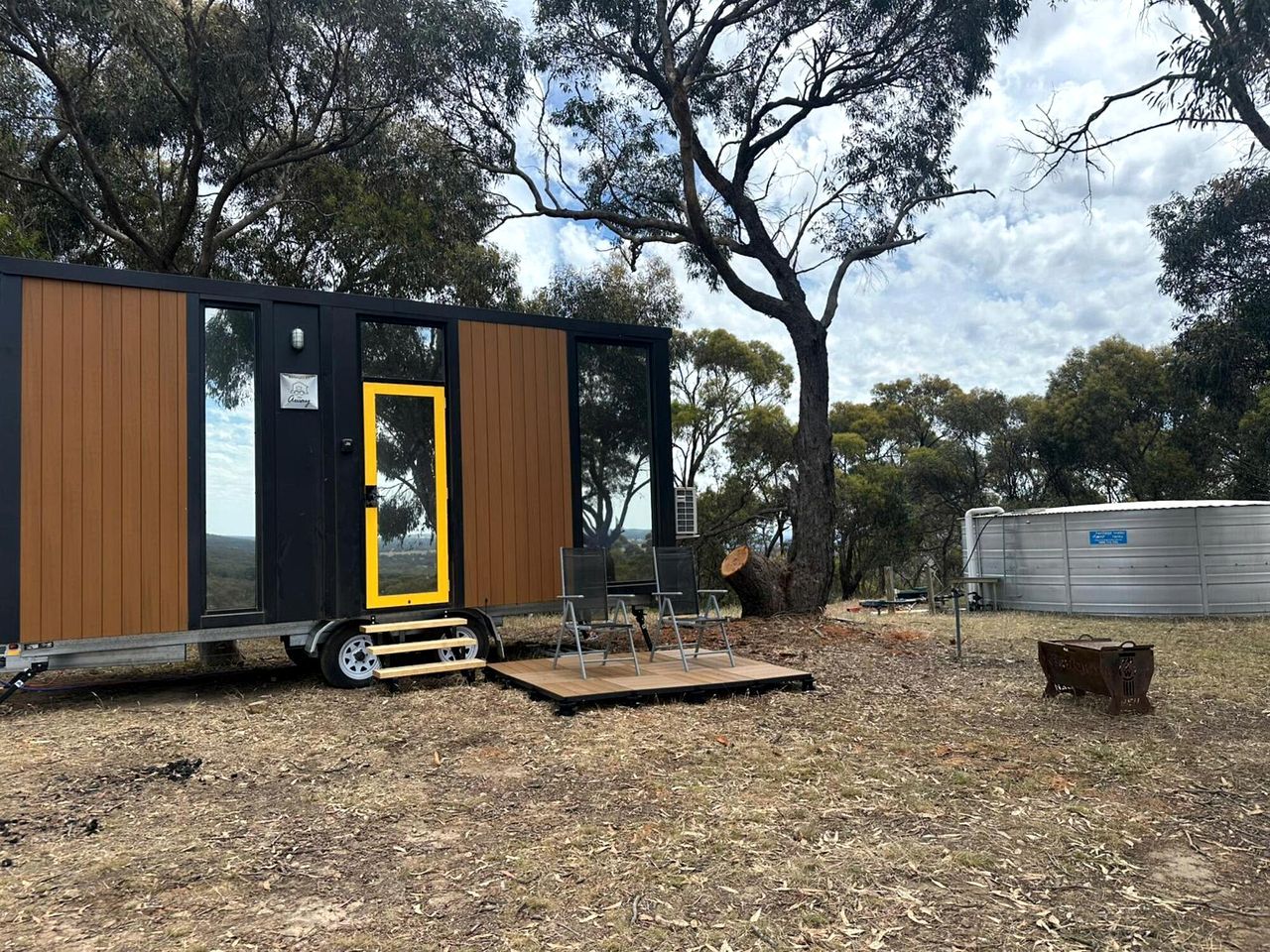 Una casa móvil moderna con fachada de madera, una puerta amarilla y un pequeño espacio exterior con sillas.