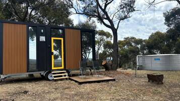 Una casa móvil moderna con fachada de madera, una puerta amarilla y un pequeño espacio exterior con sillas.