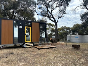 A modern mobile home with a wooden exterior, a yellow door, and a small outdoor seating area with chairs.