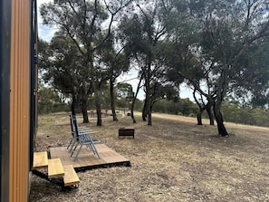 An outdoor seating area with wooden benches and metal chairs, surrounded by trees.