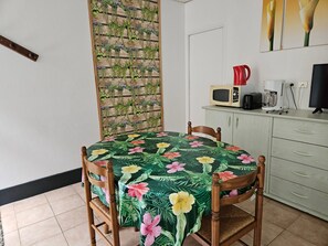 Et spiseområde med et bord dækket af en blomstret dug, træstole, en plantekasse på væggen, en mikrobølgeovn, en kaffemaskine og et fjernsyn på et skab.