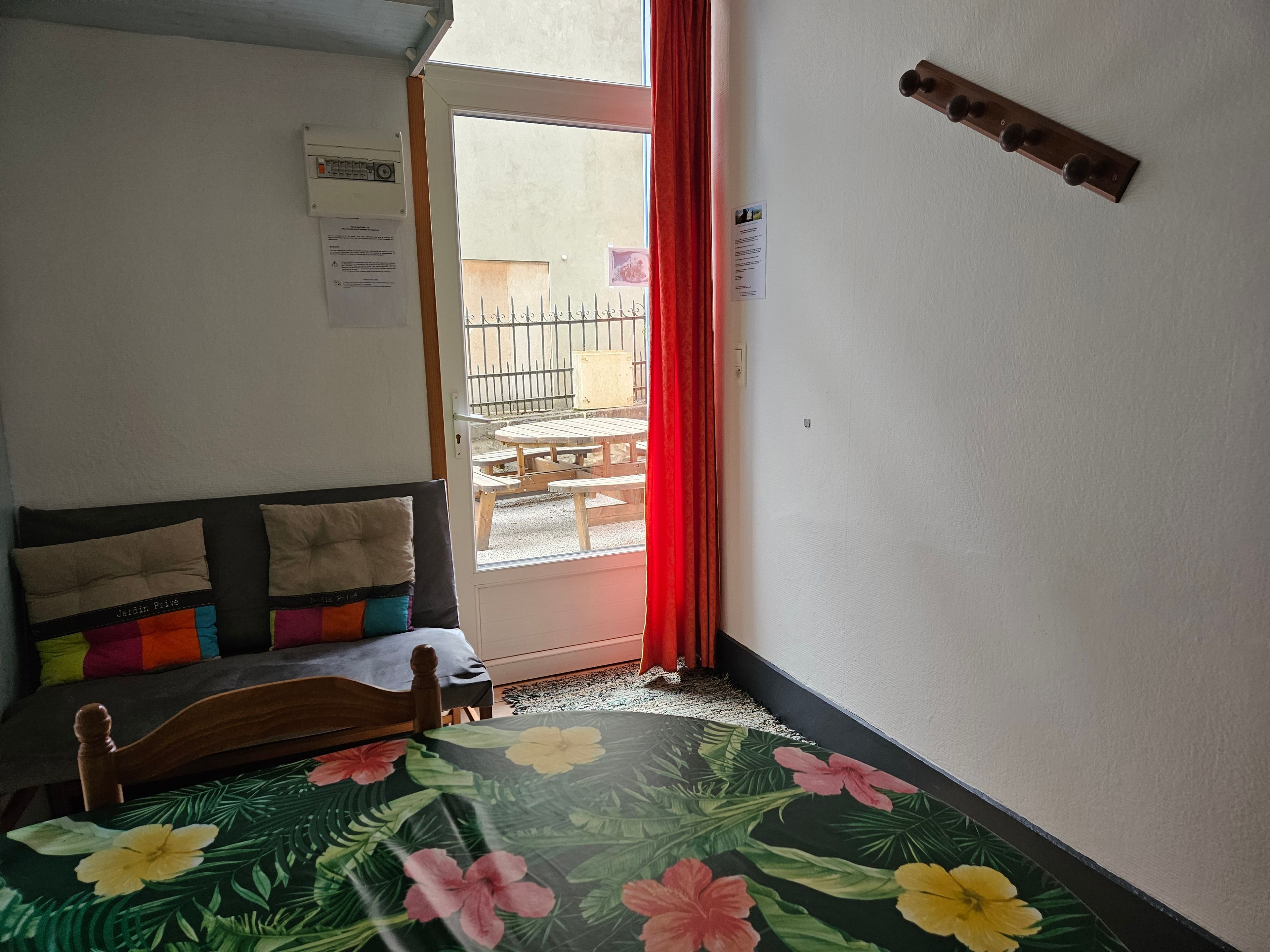 Une pièce comprenant un lit, une tête de lit en bois, une fenêtre avec un rideau rouge et une porte donnant sur un espace extérieur équipé d’une table et de chaises.