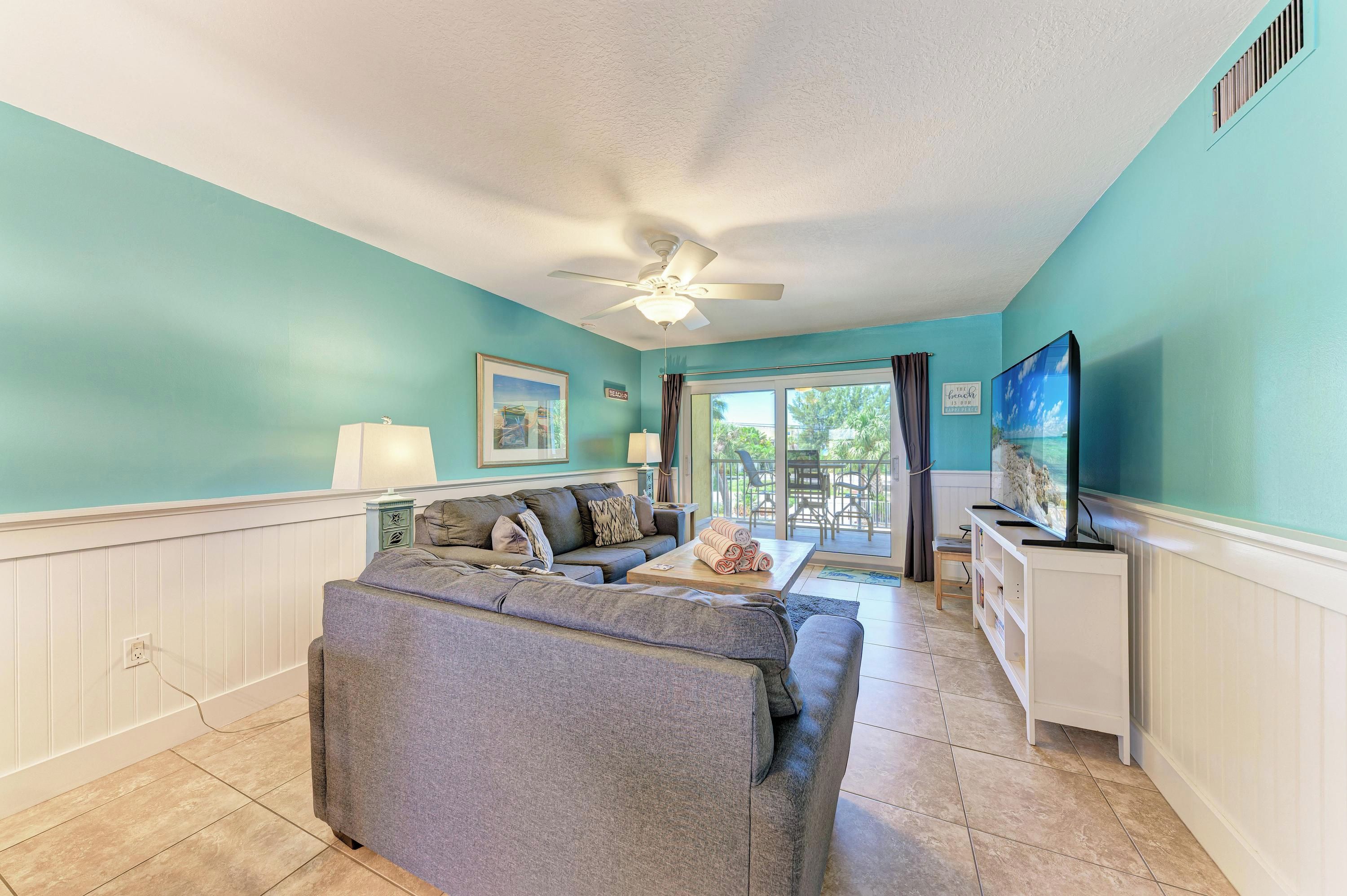 A living room with teal walls, a ceiling fan, a sofa, a coffee table, a TV, and a view of the outdoors through a sliding glass door.