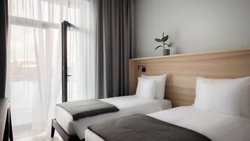 Een hotelkamer met twee bedden, een houten hoofdeinde, een kamerplant en uitzicht door een raam met transparante gordijnen.