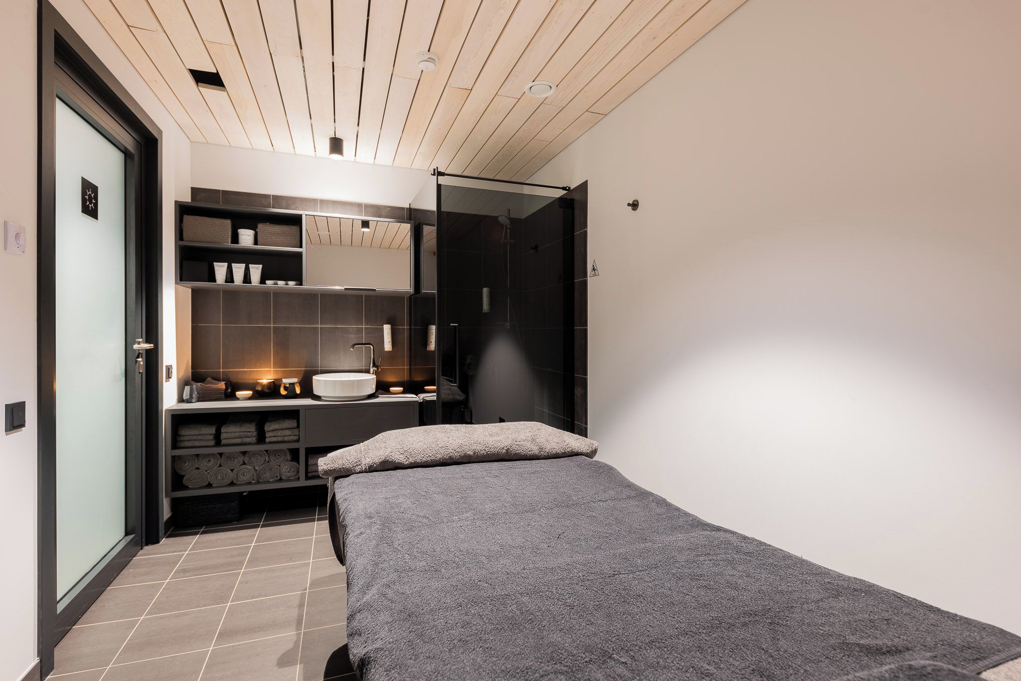 Une chambre avec un lit, une étagère intégrée où sont rangés les articles de toilette, une douche et une porte donnant sur une autre pièce.
