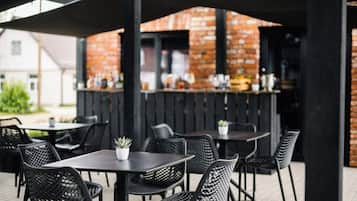 Un espace extérieur aménagé en terrasse, avec des tables et des chaises, un mur de briques en arrière-plan et un bar où divers articles sont exposés.