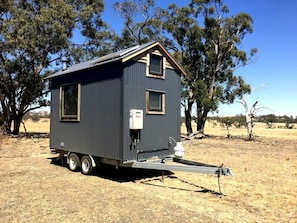 A small, dark-colored mobile home with a flat roof, a single window, and a small attached structure with a ramp.
