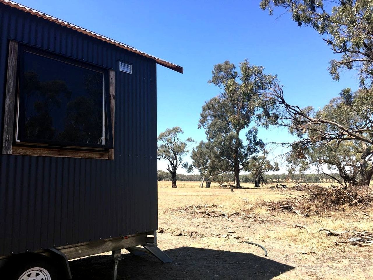 A small, dark-colored mobile home with a window and a metal roof, situated in a dry, open landscape with sparse trees.