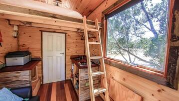 L'intérieur d'une cabane, avec un revêtement mural en bois, une échelle, une porte et une fenêtre donnant sur les arbres.