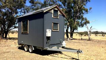 Une petite maison mobile de couleur sombre, dotée d’un toit plat, d’une seule fenêtre et d’une petite structure attenante avec une rampe d’accès.