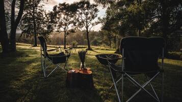 An outdoor setting with two camping chairs and a campfire, surrounded by trees and a grassy area.