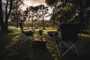 An outdoor setting with two camping chairs and a campfire, surrounded by trees and a grassy area. - Charming Tiny House Rental for Glamping in NSW (Comleroy)