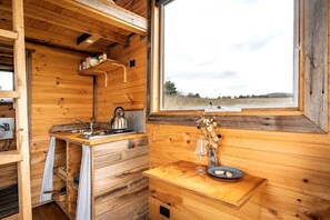 A wooden cabin interior with a kitchen area, a window with a view, and a small table with a vase and plate. - Tiny House Situated on a Brilliant Victoria Goat Farm (Monegeetta)