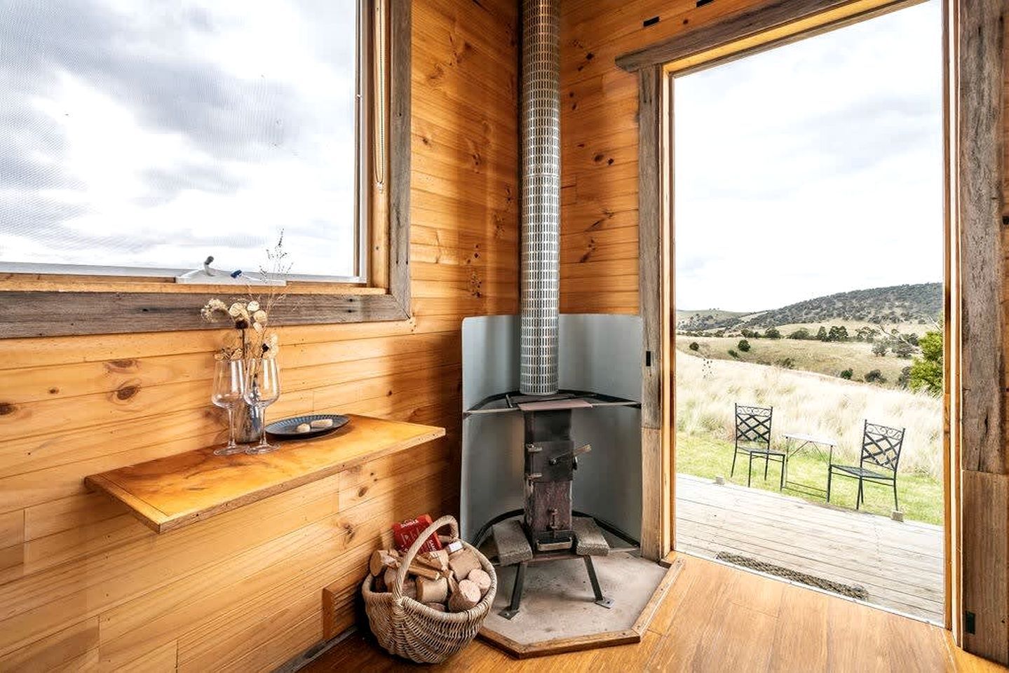 L’intérieur d’un chalet rustique, avec un poêle à bois, une fenêtre donnant sur le paysage extérieur et une petite table sur laquelle se trouvent un verre et une assiette.