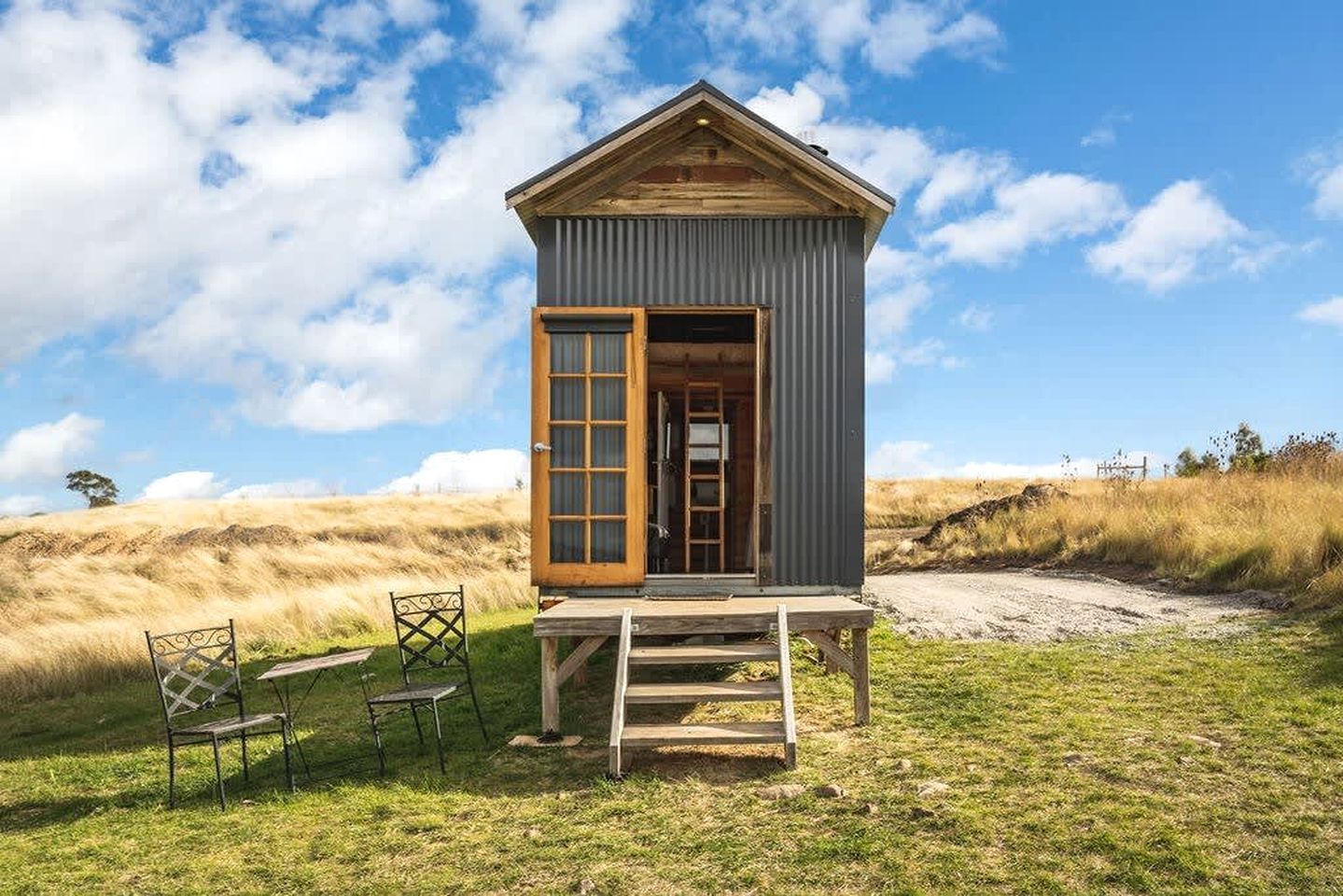 Une petite cabane en bois avec une porte ouverte, un escalier en bois et deux chaises à l’extérieur.
