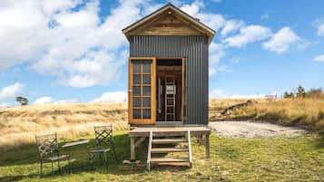 Une petite cabane en bois avec une porte ouverte, un escalier en bois et deux chaises à l’extérieur.