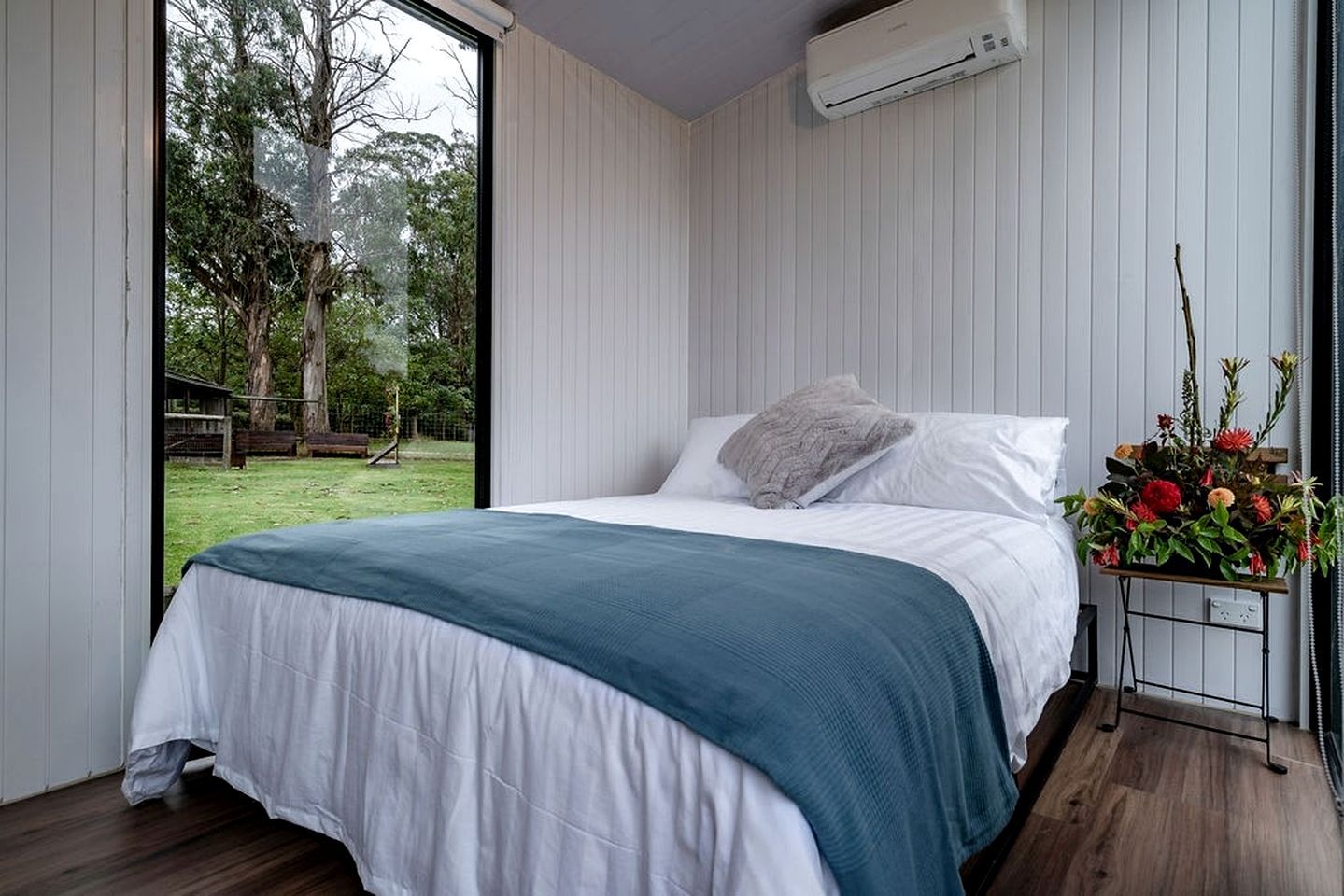 A bedroom with a bed, a window, an air conditioning unit, and a flower arrangement.