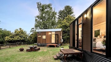 A modern cabin with a wooden exterior, a patio area with a table and benches, and a large window.